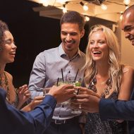 Group of young multiethnic friends enjoying evening and drinking cocktails. Happy men and women raising a toast with mojito on a patio under the light bulb wire. Elegant girls and stylish guy having fun together at party night.